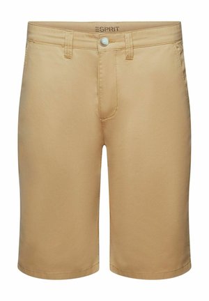 Mand iført blå denimskjorte over en stribet t-shirt, beige shorts, hvide sokker og hvide sneakers, der står mod en orange baggrund.