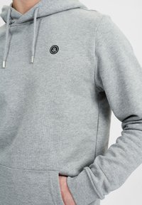 Felpa grigia con cappuccio caratterizzata da un cordino e un piccolo logo circolare. Realizzata in tessuto morbido con una superficie liscia e design a tasca a marsupio.