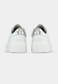 Λευκά αθλητικά παπούτσια με υφή στην λευκή σόλα, που διαθέτουν μεταλλικό ασημί ταμπελάκι στο πίσω μέρος και λείο δερμάτινο πάνω μέρος. Μινιμαλιστικός σχεδιασμός χωρίς σχέδια.