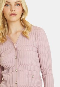 Różowy, prążkowany kardigan z dekoltem w serek, z zapięciem na guziki i perłowymi akcentami. Posiada teksturowany wzór oraz dwie funkcjonalne kieszenie z przodu.