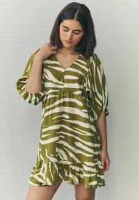 Kurzarmkleid in olivgrün mit weißen Zebra-Streifen, V-Ausschnitt und ausgestelltem, gerafftem Saum. Aus leichtem Stoff gefertigt.