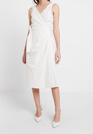 Femme portant une robe portefeuille blanche sans manches avec un lien sur le côté et des talons blancs à bouts pointus, debout devant un fond uni.