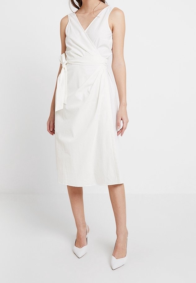 Femme portant une robe portefeuille blanche sans manches avec un lien sur le côté et des talons blancs à bouts pointus, debout devant un fond uni.