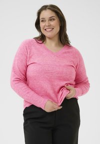 Ružový sveter s V-výstrihom a dlhými rukávmi, vyrobený z mäkkej zmesi látok. Mierne textúrovaný, s voľným strihom, skombinovaný s čiernymi nohavicami.
