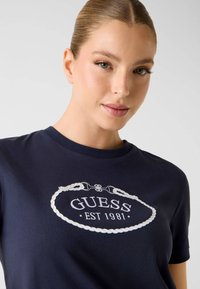Granatowy bawełniany t-shirt z białym haftowanym logo „GUESS EST 1981” wewnątrz splecionej ramki. Klasyczny krój z okrągłym dekoltem.