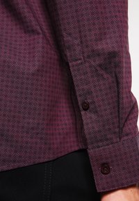 Chemise à manches courtes de couleur bourgogne profond avec un motif circulaire subtil. Dotée d'un ourlet arrondi et de boutons foncés aux poignets. Texture lisse.