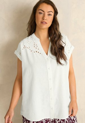 Kvinde med langt brunt hår iført en hvid skjorte med knapper og blonder med hulmønster på skuldrene samt opkrogede korte ærmer.