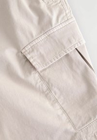 Beige színű vászonsort, sima textúrával, oldalsó zsebből álló zsebbel, és látható varrási részletekkel a széleken.