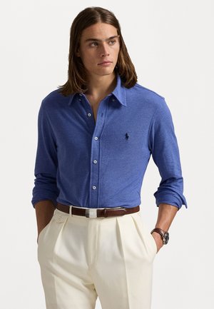 Blue long-sleeve polo shirt with a buttoned collar, featuring a small embroidered logo on the chest, paired with cream pleated trousers.