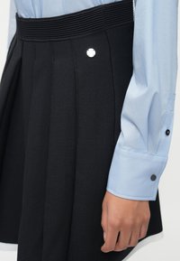 Čierna plisovaná sukňa s textúrovaným povrchom a strieborným gombíkom, skombinovaná so svetlomodrou košeľou s manžetami na gombíky.