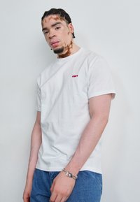 T-shirt branca de algodão com mangas curtas e decote redondo. Apresenta um logótipo vermelho "OBEY" na área do peito esquerdo. Usada com jeans de ganga azuis.