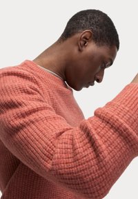 Suéter de ribete coral con cuello redondo, tela texturizada y mangas largas, mostrado en un modelo que inclina la cabeza hacia abajo.