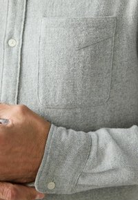 Pilkos mygtukinės marškiniai, pagaminti iš tekstūruotos medžiagos, su krūtinės kišene, standartiniais rankogaliais su mygtuko uždarymu ir audimu raštu.