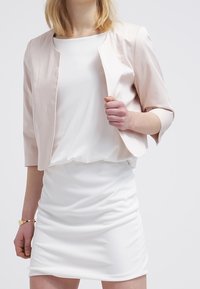 Lichtroze cropped jas met driekwart mouwen, gecombineerd met een witte nauwsluitende jurk, die gladde stoffen en een eenvoudig ontwerp laat zien.