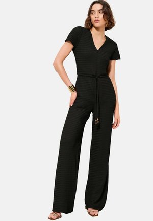 Frau trägt einen schwarzen, kurzärmeligen Overall mit V-Ausschnitt, gebundener Taille, weitem Bein, goldenem Armreif und braunen Sandalen.