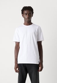 Bílé bavlněné tričko s krátkým rukávem a kulatým výstřihem. Obsahuje malé modré logo blízko lemu. Model má na sobě černé džíny.