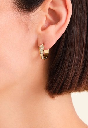 Orecchino a cerchio in oro con un design ondulato, caratterizzato da una fila di piccoli cristalli chiari su un lato, posizionato su capelli corti e scuri.