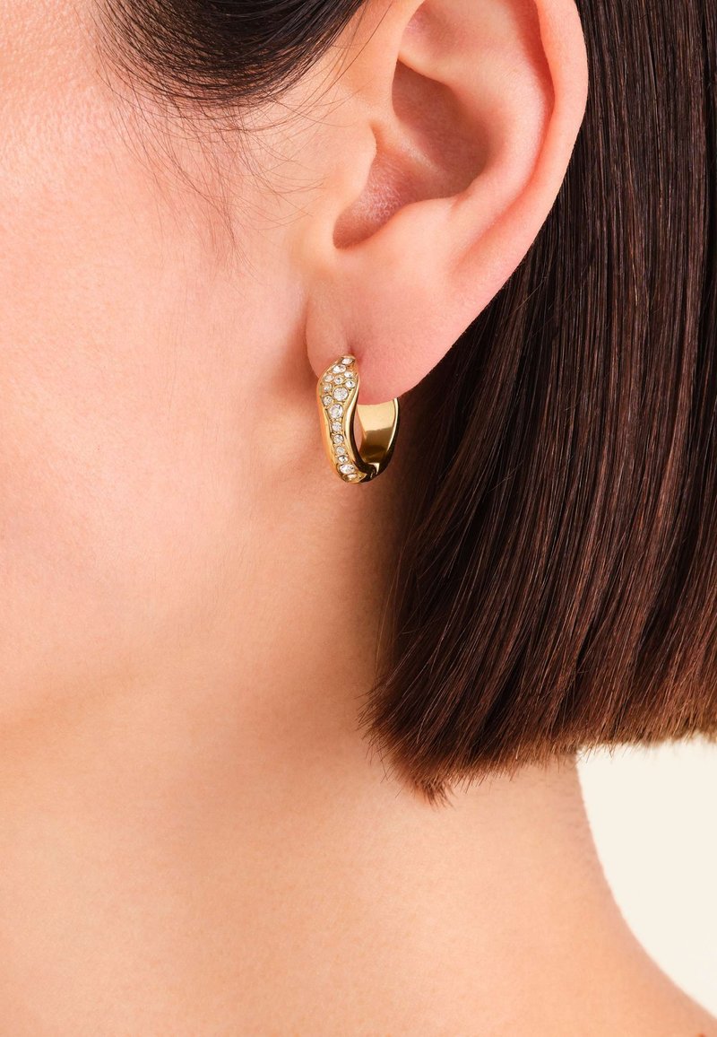 Pendiente de aro dorado con un diseño ondulado, que presenta una fila de pequeños cristales transparentes a lo largo de un lado, sobre cabello corto y oscuro.