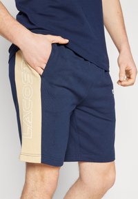 Shorts en coton bleu marine avec un panneau latéral beige portant le logo "LACOSTE" en blanc, taille élastique et poche latérale.