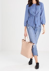 Niebieska bluzka w paski z wiązaniem z przodu, zestawiona z jasnoniebieskimi, podartymi dżinsami. Modelka nosi beżową torbę typu tote i czarne klapki mule.