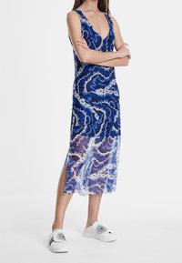Robe sans manches à motifs bleus avec un décolleté en V profond, en tissu transparent, fente sur les côtés, portée avec des chaussures blanches à enfiler ornées de motifs floraux.