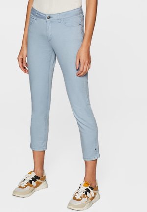 Person iført lyseblå korte bukser og hvide, sorte og tanfarvede sneakers, stående mod en ensfarvet baggrund.