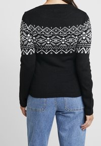 Schwarzer Pullover mit weißem Strickmuster und geometrischen Designs. Lange Ärmel und ein rundes Halsband, strukturiertes Gewebe. Blaue Jeans sind darunter sichtbar.