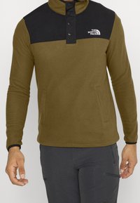 Forro polar verde oliva con panel negro en los hombros, cremallera de cuarto y broches de presión. Cuenta con bolsillos laterales y puños negros a contraste.