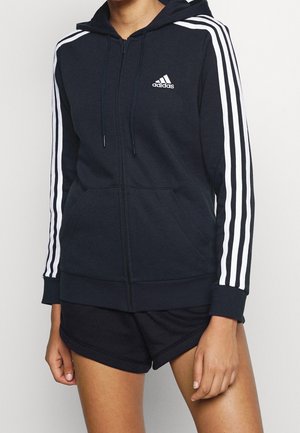 Person iført en sort Adidas hættetrøje med lynlås med hvide striber på ærmerne og sorte shorts, stående mod en neutral baggrund.