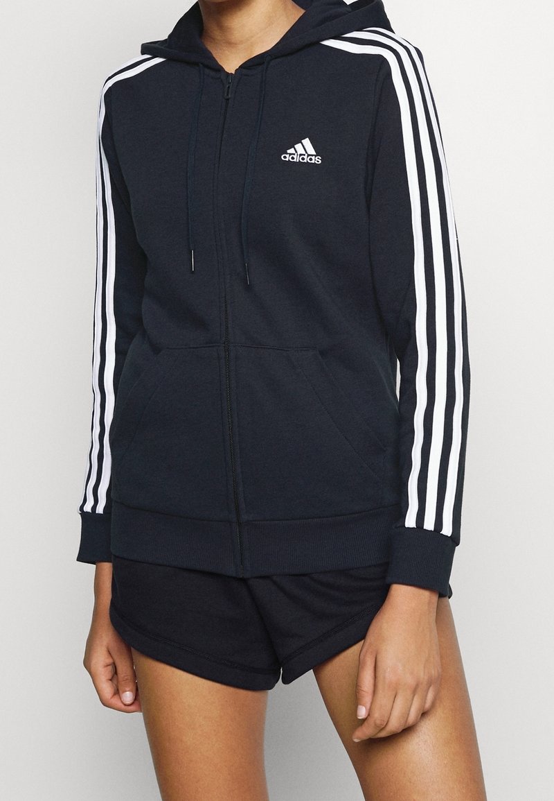 Personne portant un sweat à capuche zippé noir Adidas avec des bandes blanches sur les manches et un short noir, debout devant un fond neutre.