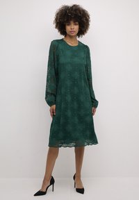 Grön spetsklänning med långa ärmar, blommönster, scallopad fåll. Bärs med svarta högklackade skor. Slät textur och avslappnad passform.