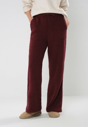 Person iført løse vinrøde fleecebukser med lommer, beige casual sko og cremefarvet striktrøje, stående mod en ensfarvet lys baggrund.