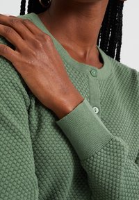 Green knitted cardigan featuring a textured pattern, round neckline, and button closure. Ribbed cuffs and a soft, lightweight fabric.