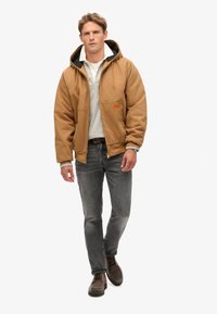 Chaqueta aislante marrón con capucha, suéter gris debajo, pantalones vaqueros grises y zapatos marrones. Cuenta con dos bolsillos frontales y una cremallera.