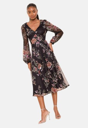 Naine kannab musta midi pikkusega kleiti, millel on roosade ja oranžide lillepistega tikand ning läbipaistvad pikad varrukad, kombineeritud rihmjalatsitega kõrge kontsaga sandaalides.