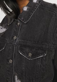 Detailní záběr na osobu ve černé roztrhané džínové bundě s kovovými cvoky a předními kapsami na klopu.