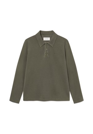 Ung person med langt rødt hår iført en grøn langærmet skjorte, løse mørke jeans og sorte sko, står med hænderne i lommerne.