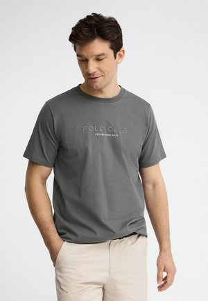 Mężczyzna w szarej koszulce "Polo Club" i beżowych spodniach, z rękami w kieszeniach, patrzący w dół z neutralnym wyrazem twarzy na jednolitym tle.