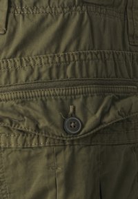 Olivgrön tyg med strukturerad yta, synliga stygn, en knappstängning och en ficka med dragkedja på den nedre delen.