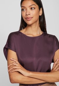 Γυναίκα με μακριά καστανά μαλλιά, φορώντας ένα μοβ σατέν κοντομάνικο μπλουζάκι, με τα χέρια σταυρωμένα, κοιτάζοντας προς τα πλάγια με φόντο ένα απαλό ανοιχτό χρώμα.