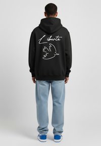 Sweat à capuche noir avec le texte "Liberté" en blanc et un graphique de colombe au dos, associé à un jean baggy bleu clair et des baskets bleues.