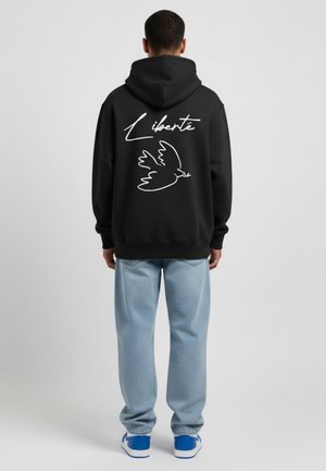 Felpa nera con testo bianco "Liberté" e grafica di una colomba sulla schiena, abbinata a jeans baggy blu chiaro e sneakers blu.