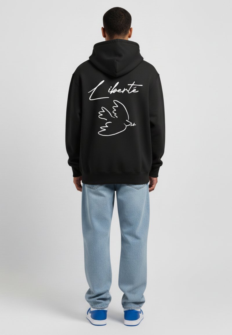 Sweat à capuche noir avec le texte "Liberté" en blanc et un graphique de colombe au dos, associé à un jean baggy bleu clair et des baskets bleues.