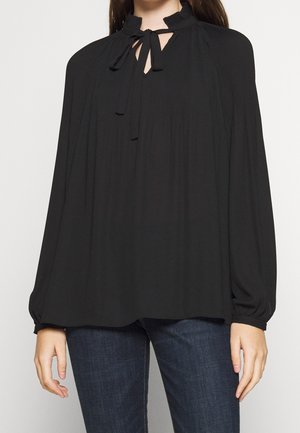 Femme portant un chemisier noir ample avec un col montant à volants, des manches longues, et un détail à nouer au col, associé à un jean bleu foncé.