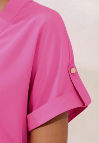 Blusa a maniche corte di un luminoso rosa, con una texture morbida e liscia, polsini arrotolati e dettagli in bottone dorato sulle maniche.