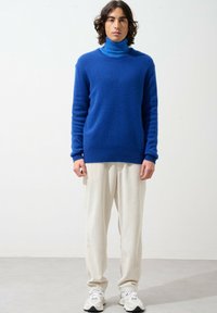 Blauer gestrickter Rollkragenpullover mit gerippten Bündchen und Saum, kombiniert mit cremefarbenen texturierten Hosen und leichten Sneakers.