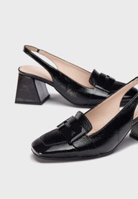Des chaussures slingback en cuir verni noir avec un bout carré, des talons blocs et un accent de boucle décorative sur le devant; texture lisse.