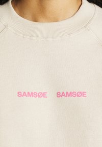 Sweatshirt beige avec un col côtelé et deux inscriptions du nom de la marque "SAMSØE" imprimées en rose. Texture de tissu doux visible.