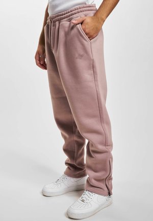 Lys pink sweatpants lavet af blød stof med en elastisk talje, sidelommer og lynlåsdetailler ved anklerne, parret med hvide sneakers.