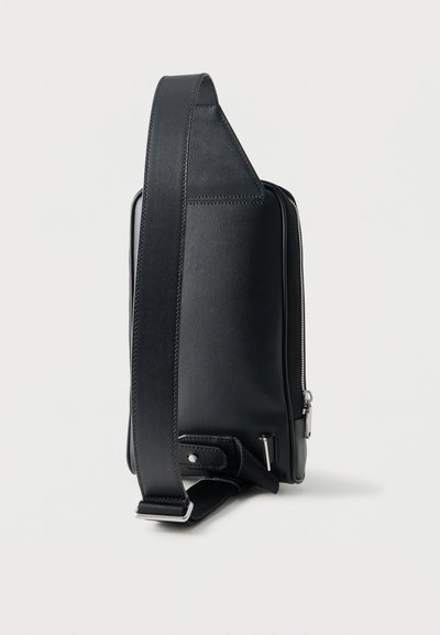 Sac bandoulière compact noir avec sangle large réglable, fermeture éclair argentée et détails de couture minimalistes sur fond blanc uni.